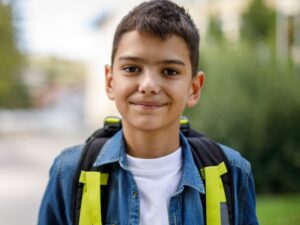 صبي مراهق مبتسم يحمل حقيبة مدرسية يقف أمام مدرسته.