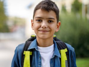 Smiling teenage boy with a school bag is standing in front of his school.