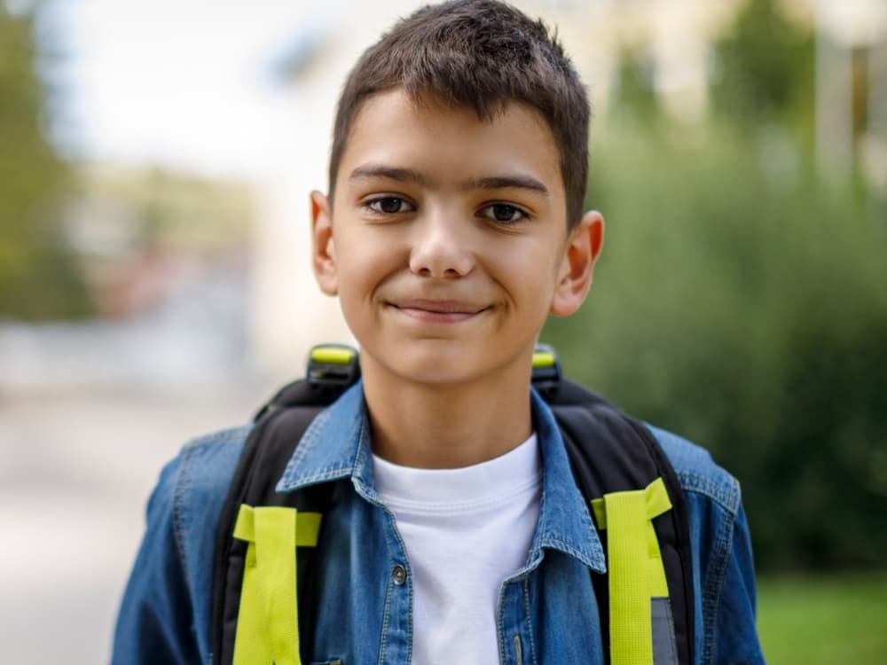 Smiling teenage boy with a school bag is standing in front of his school.