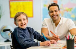 A young boy in a wheelchair works with his teacher in a classroom. They are both smiling.