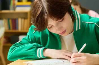 A teenage student sits at a desk in the classroom. They are focused and writing notes in a book.