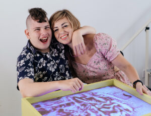 A teenage boy with disability and a Mohican style haircut sits with his arm around his mother's shoulder. They are both smiling and doing a sand puzzle.