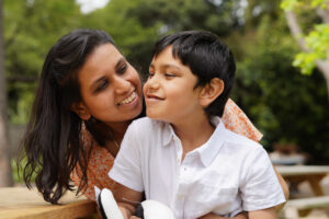 A young boy with disability is in the park with his Mum. He smiles and looks into the distance and she looks over his shoulder and smiles at her son.