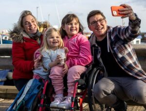 A family of four are outdoors. The father is holding a mobile phone to take a selfie of the group. The child in the center of the group is in a wheelchair.