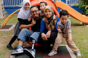 عائلة مسلمة لديها ثلاثة أطفال تجلس في أسفل منزلق نفق. ترتدي الأم وابنتها الحجاب. كلهم يبتسمون.