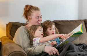 A mother sits on a sofa with her two young children on her lap. They are reading a picture book together.