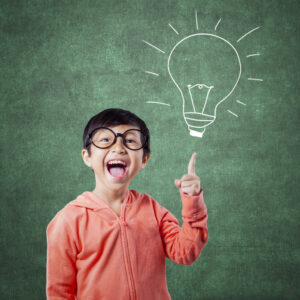 A boy with glasses and an orange top stands in front of a chalkboard. He's pointing to a drawing of a lightbulb and smiling.