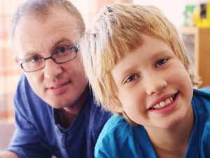 A father lays on the floor with his teenage son. He wears glasses, and they smile. The father holds an iPad.